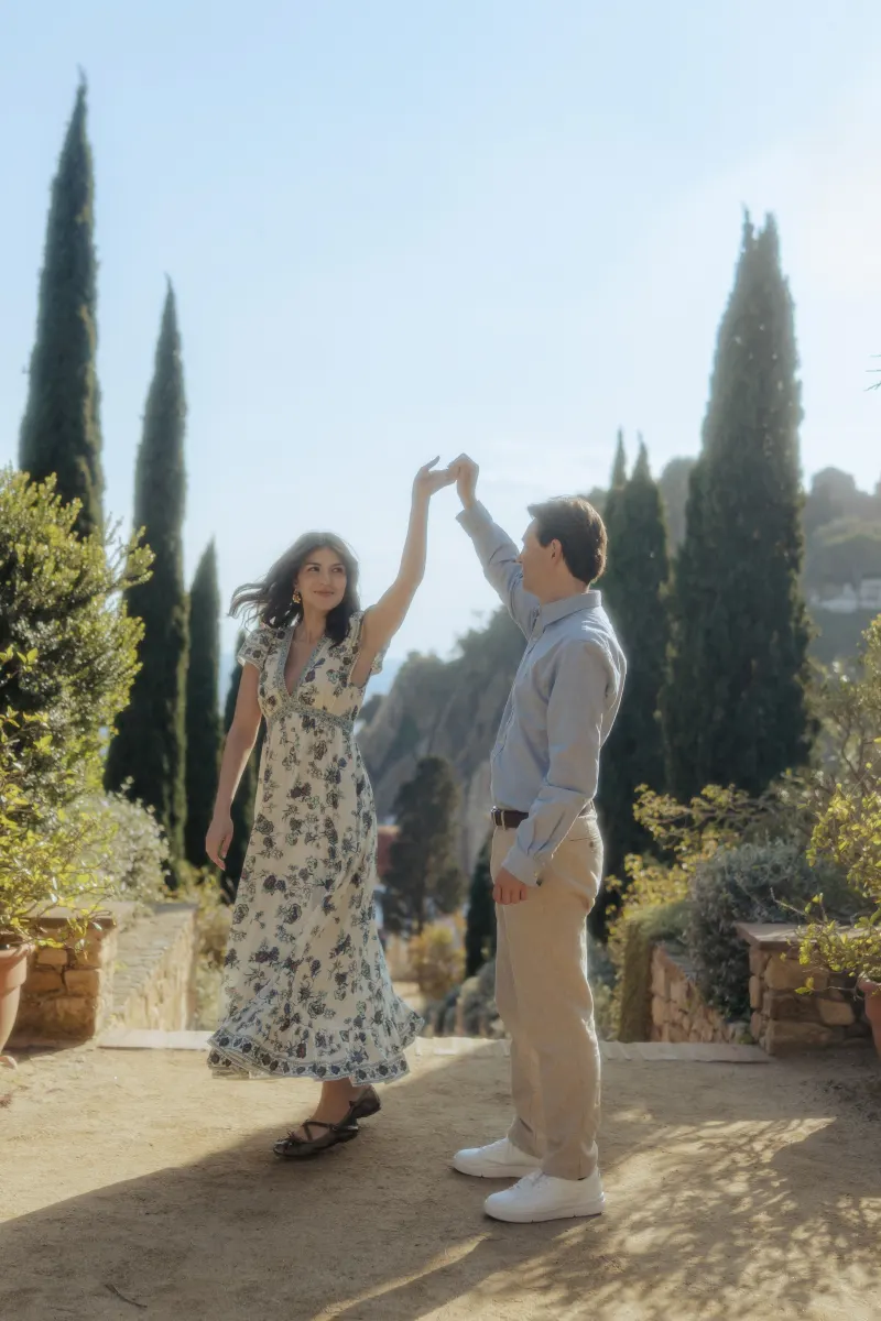 Man twirls his fiancée in a sunlit garden path, capturing a candid moment from a surprise proposal photoshoot.