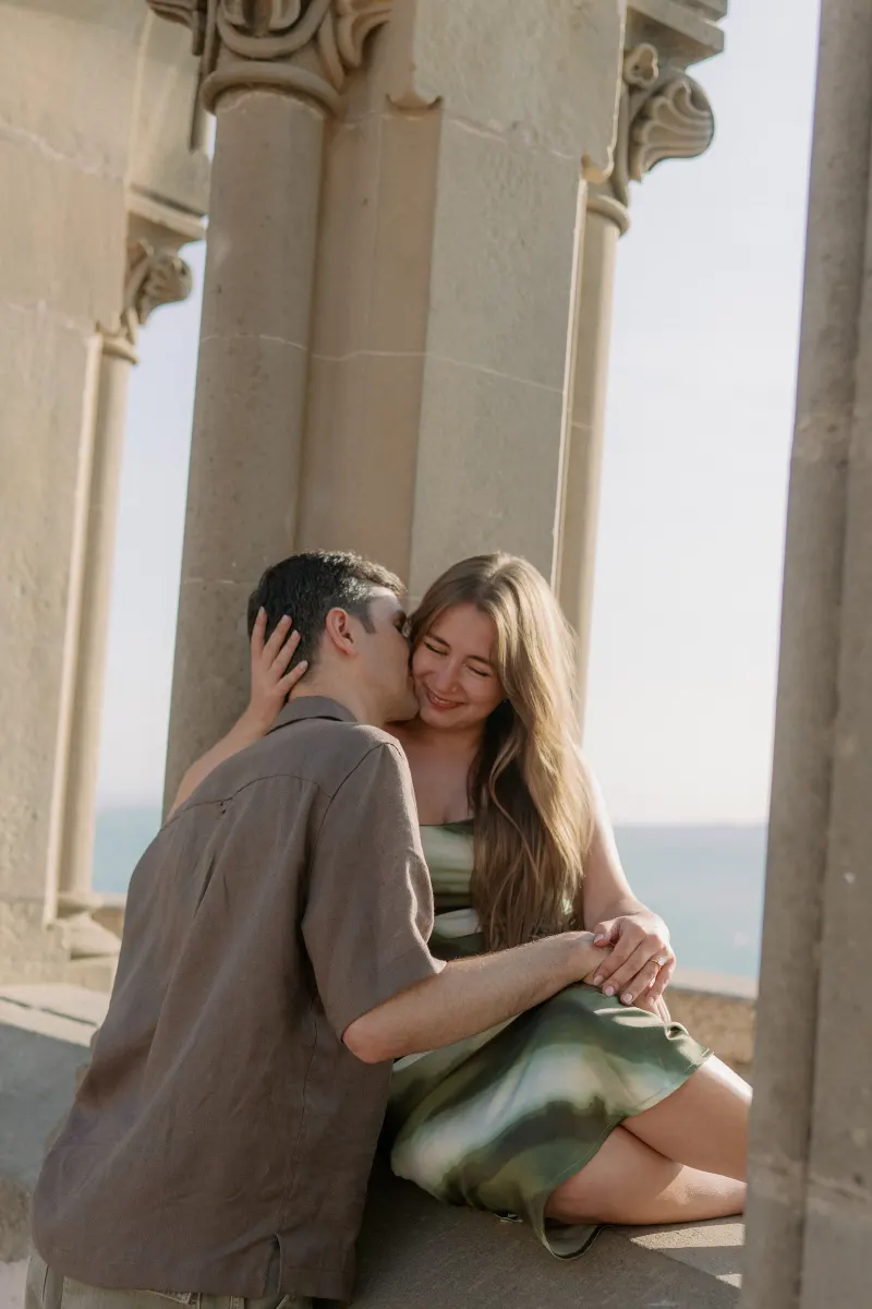 Megan rests her hand against Tim's head beside a carved stone column.