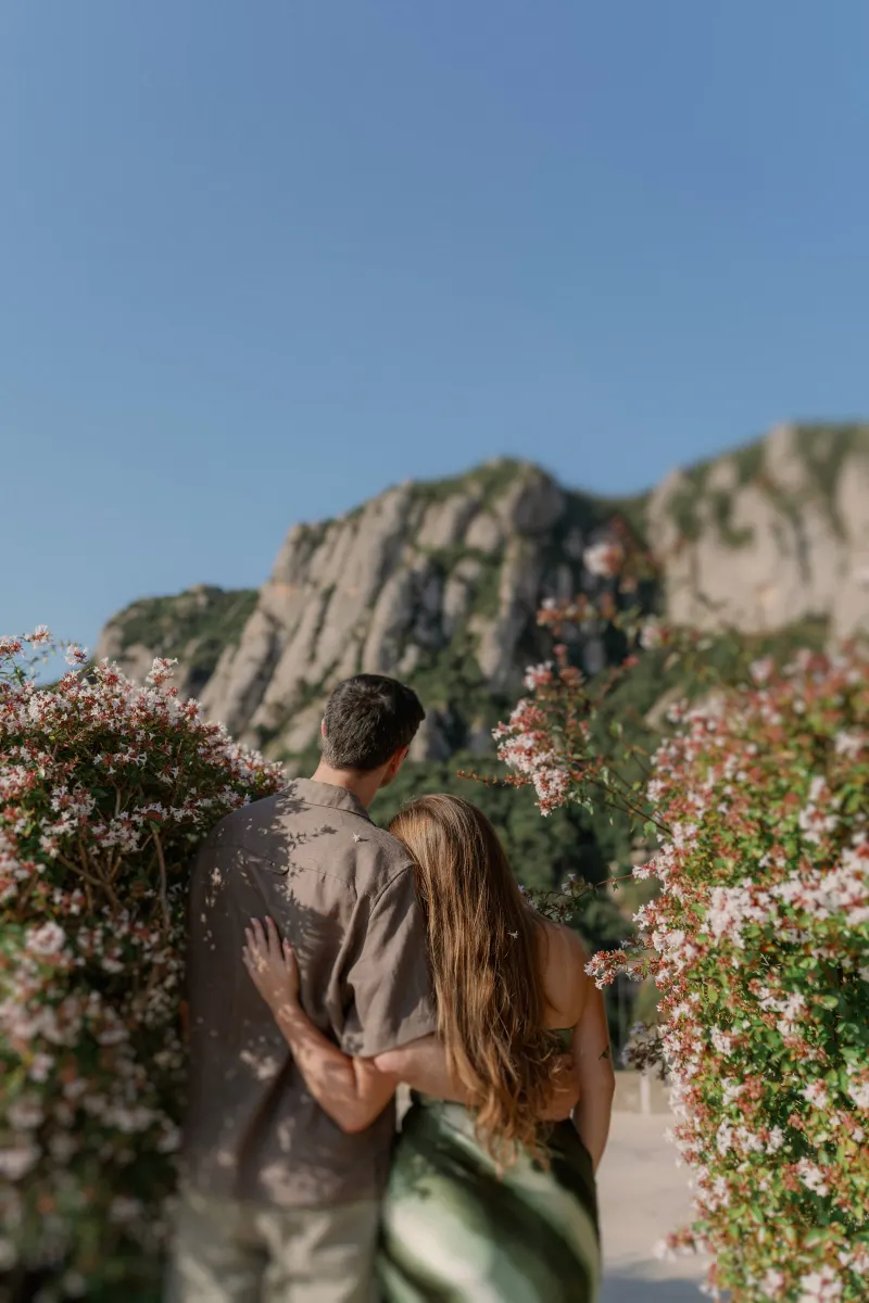 Megan wraps her arm around Tim beside flowering shrubs.