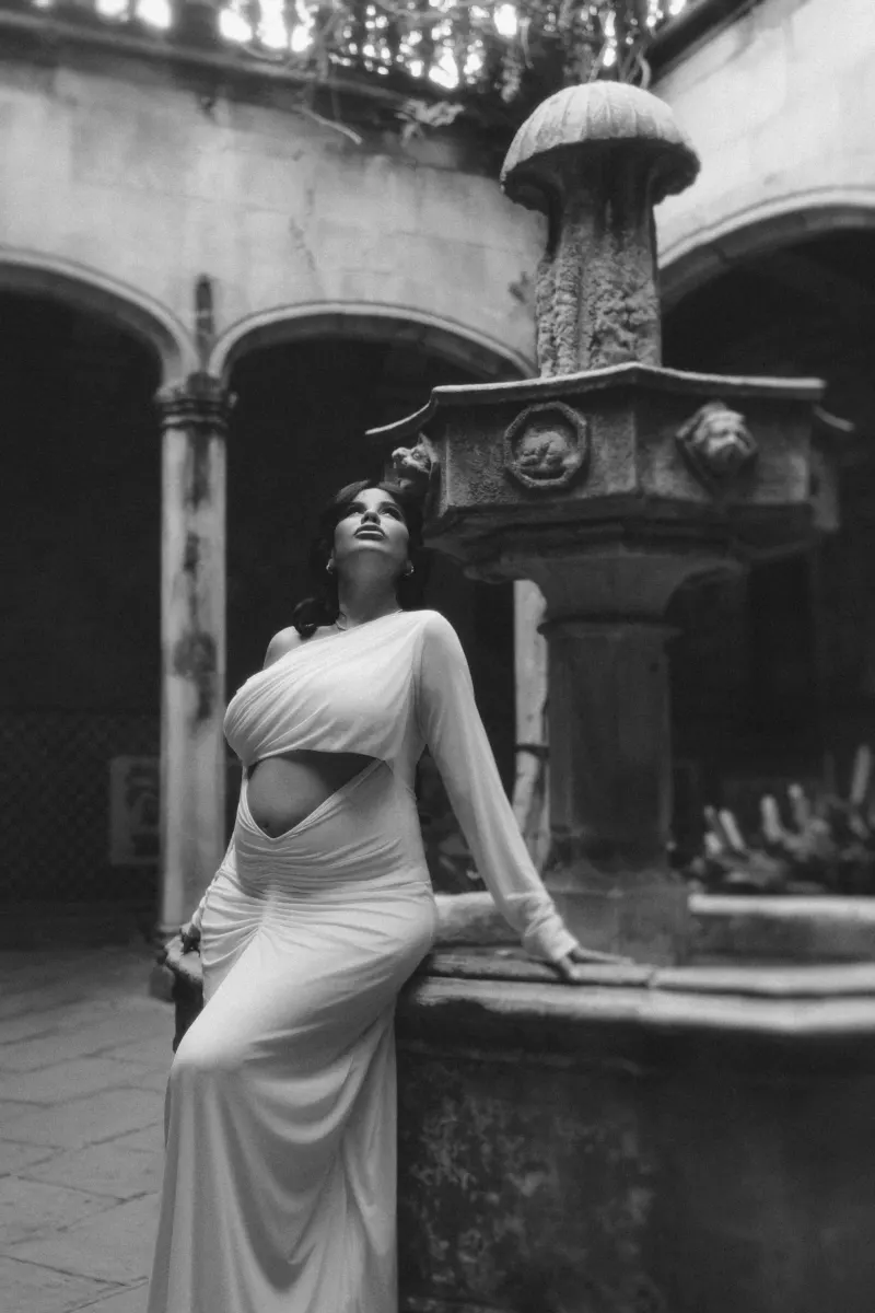 Mom-to-be in a draped white dress gazes upward beside an old fountain during a Vacation Portraits shoot in a courtyard.