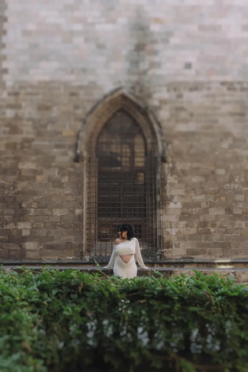 Mom-to-be in a white dress poses serenely by lush greenery during a Vacation Portraits photoshoot in a historic courtyard.