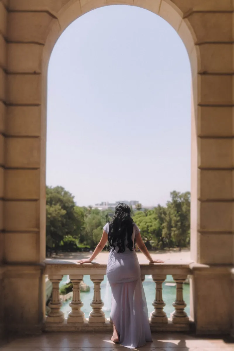 Mother-to-be in a lilac dress gazes over a sunlit park from a grand balcony, Vacation Portraits in Barcelona.