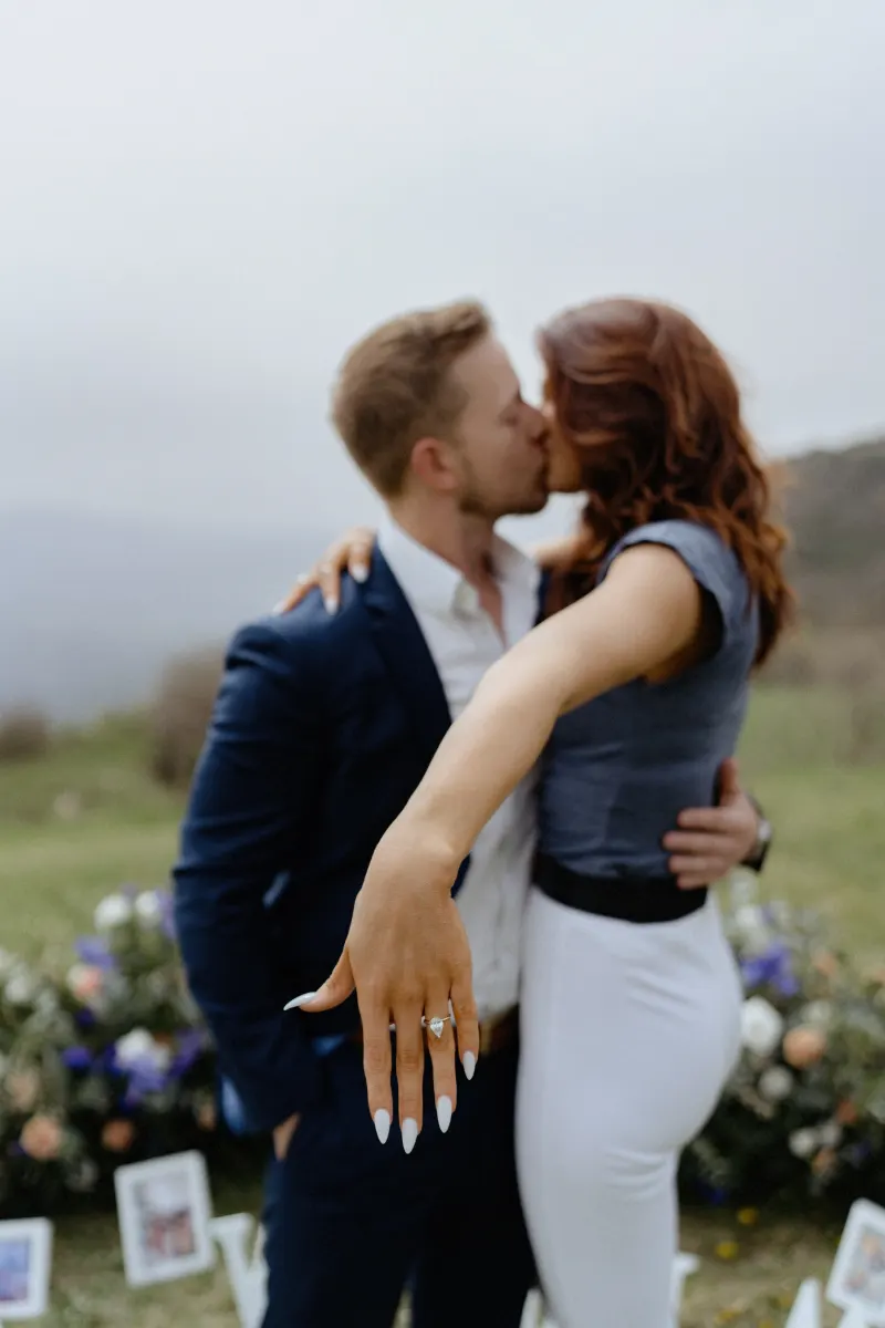Newly engaged couple kisses on a hillside, with the bride-to-be showing off her ring during a surprise proposal photoshoot.