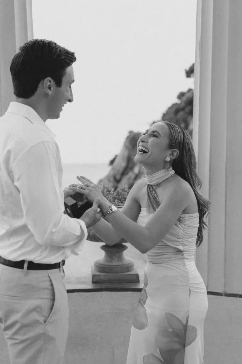 Overjoyed woman laughs as her partner proposes at a scenic seaside pavilion during a surprise proposal photoshoot.