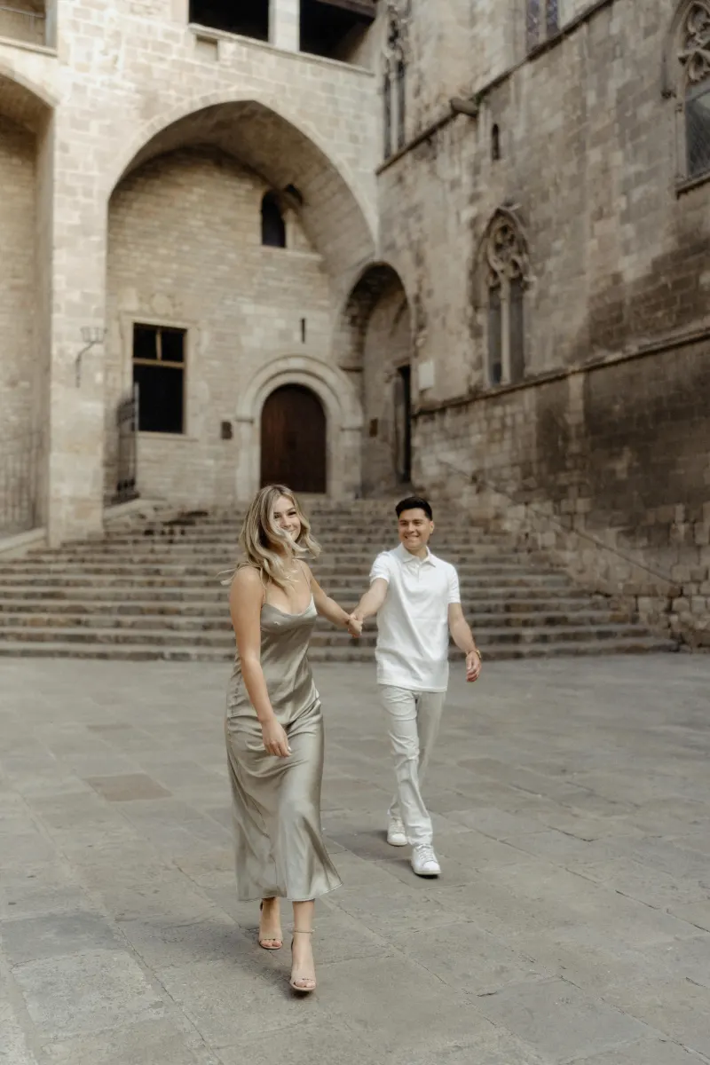 Playful couple holding hands and smiling while exploring a historic Barcelona courtyard during a Vacation Portraits photoshoo