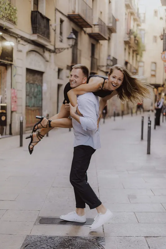 Playful couple laughing as he carries her over his shoulder down a charming city street during an engagement photoshoot.