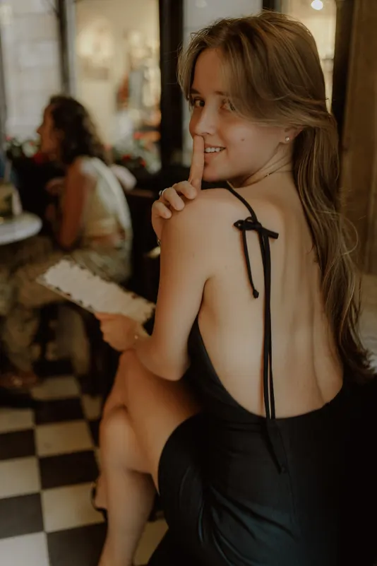 Playful woman in a black dress smiles with a shushing gesture during a Vacation Portraits photoshoot at a cozy café.