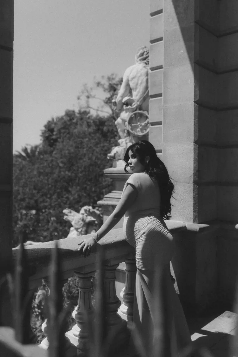 Pregnant woman gazes thoughtfully from a sunlit balcony, Vacation Portraits photoshoot in Barcelona.