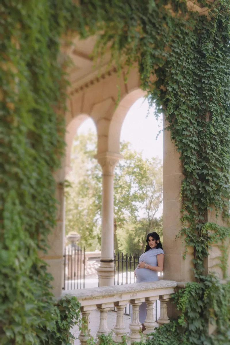 Pregnant woman in a blue dress leans on a vine-covered balcony, Vacation Portraits photoshoot in Barcelona.