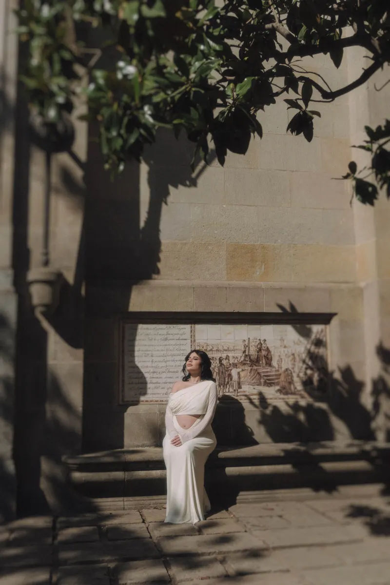 Pregnant woman in a flowing white dress sits peacefully on a sunlit stone bench during a Vacation Portraits session.
