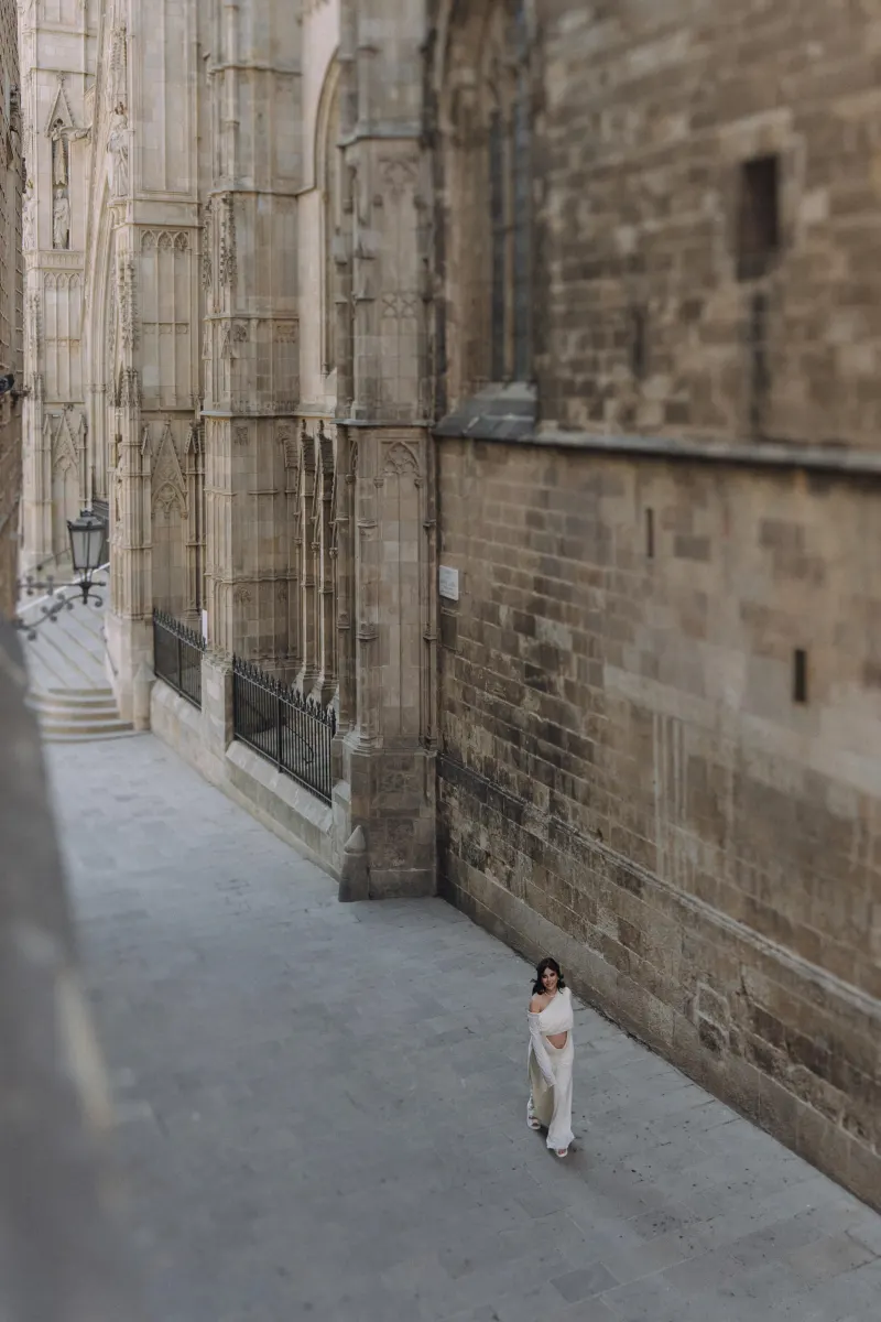 Pregnant woman in a flowing white dress strolls alone along a historic stone street during a Vacation Portraits photoshoot.