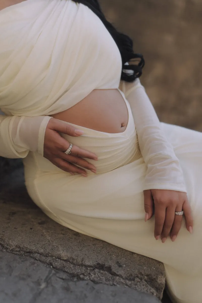Pregnant woman in a soft white gown gently holds her belly, captured during a Vacation Portraits maternity session.