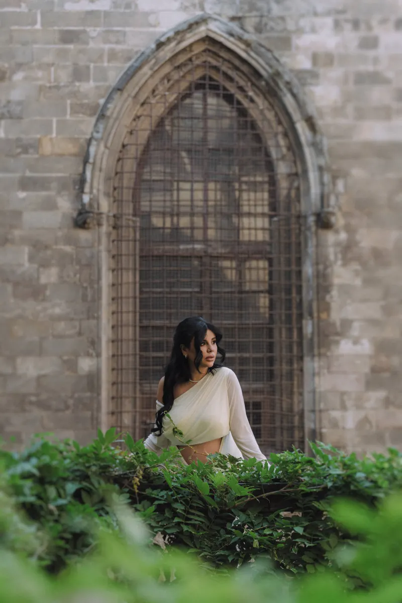 Pregnant woman in a white dress leans on a garden wall, framed by greenery during a Vacation Portraits photoshoot.