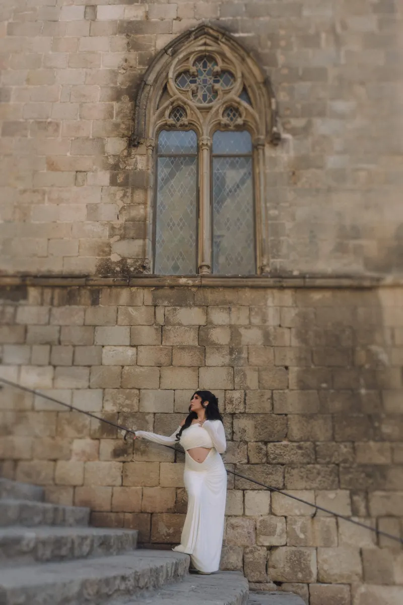 Pregnant woman in a white dress pauses on stone steps beneath a gothic window during a Vacation Portraits session.