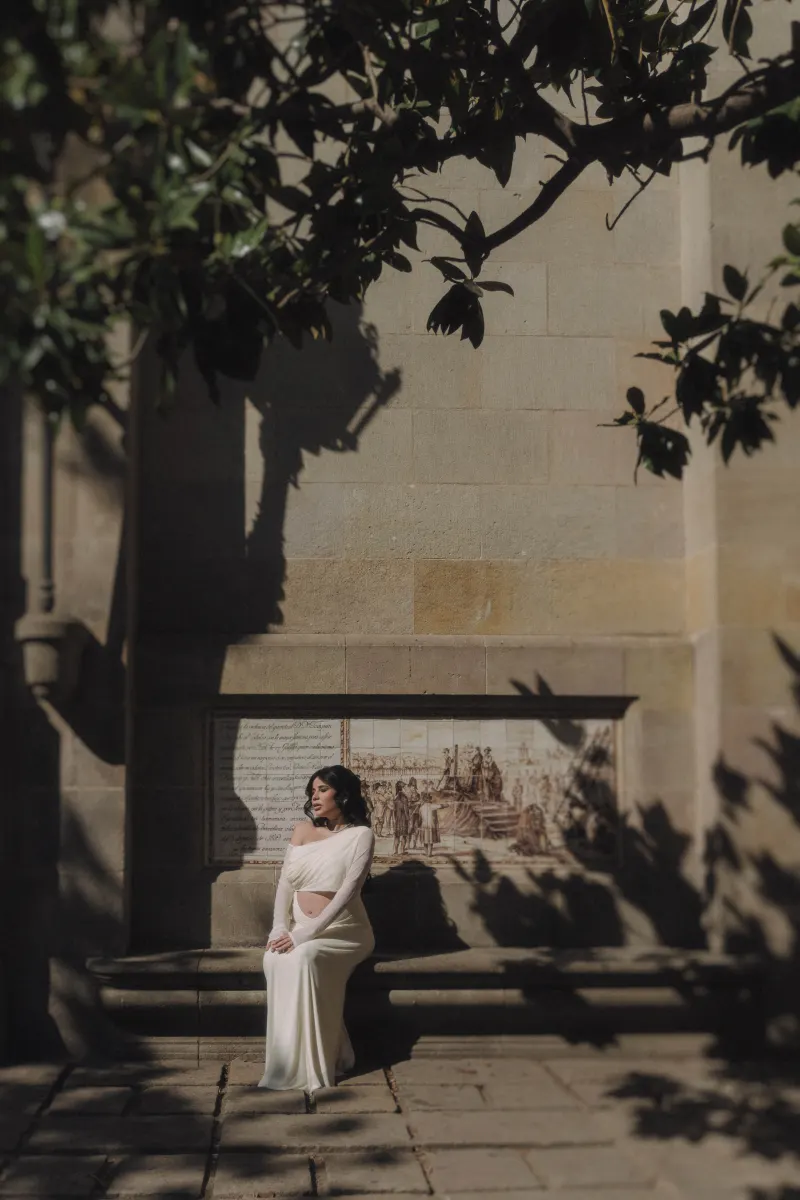 Pregnant woman in a white dress sits serenely on a sunlit stone bench during a Vacation Portraits session in the city.
