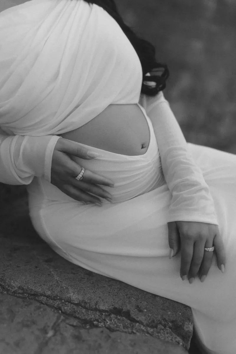 Pregnant woman in a white gown gently holds her belly, captured during a Vacation Portraits session outdoors.