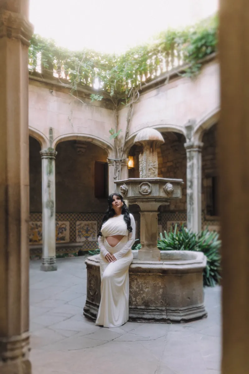 Pregnant woman in a white gown poses serenely by a stone fountain during a Vacation Portraits shoot in a historic courtyard.