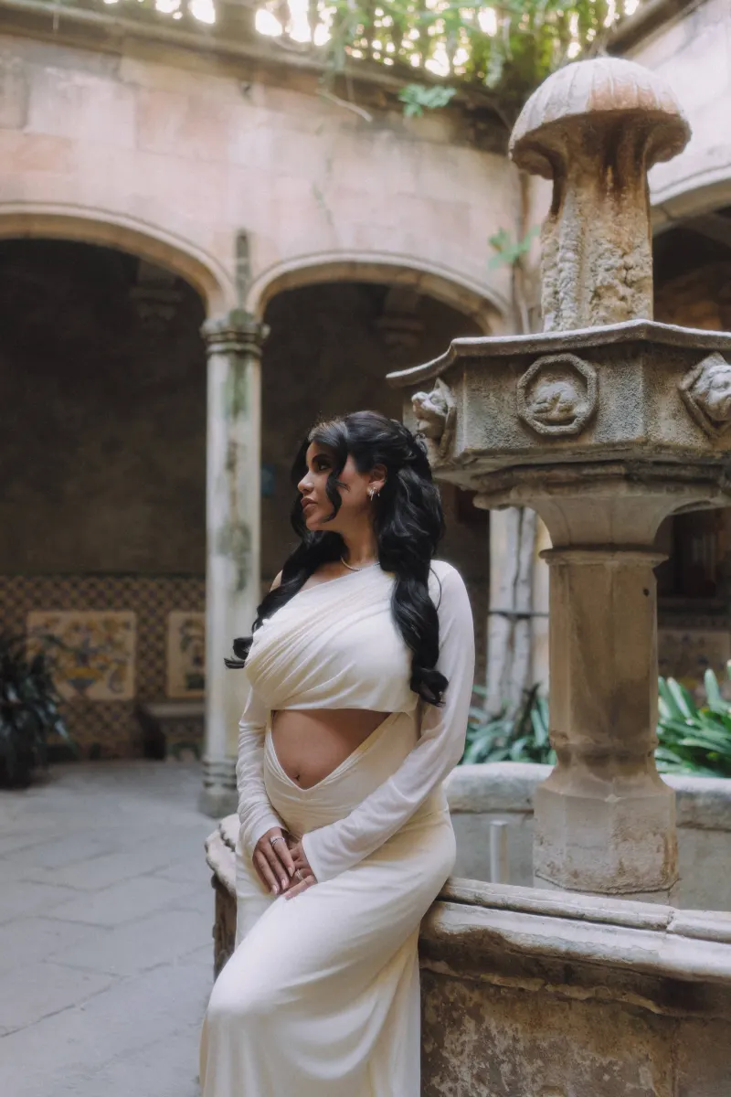 Pregnant woman in a white gown rests by a historic stone fountain during a Vacation Portraits session in a sunlit courtyard.