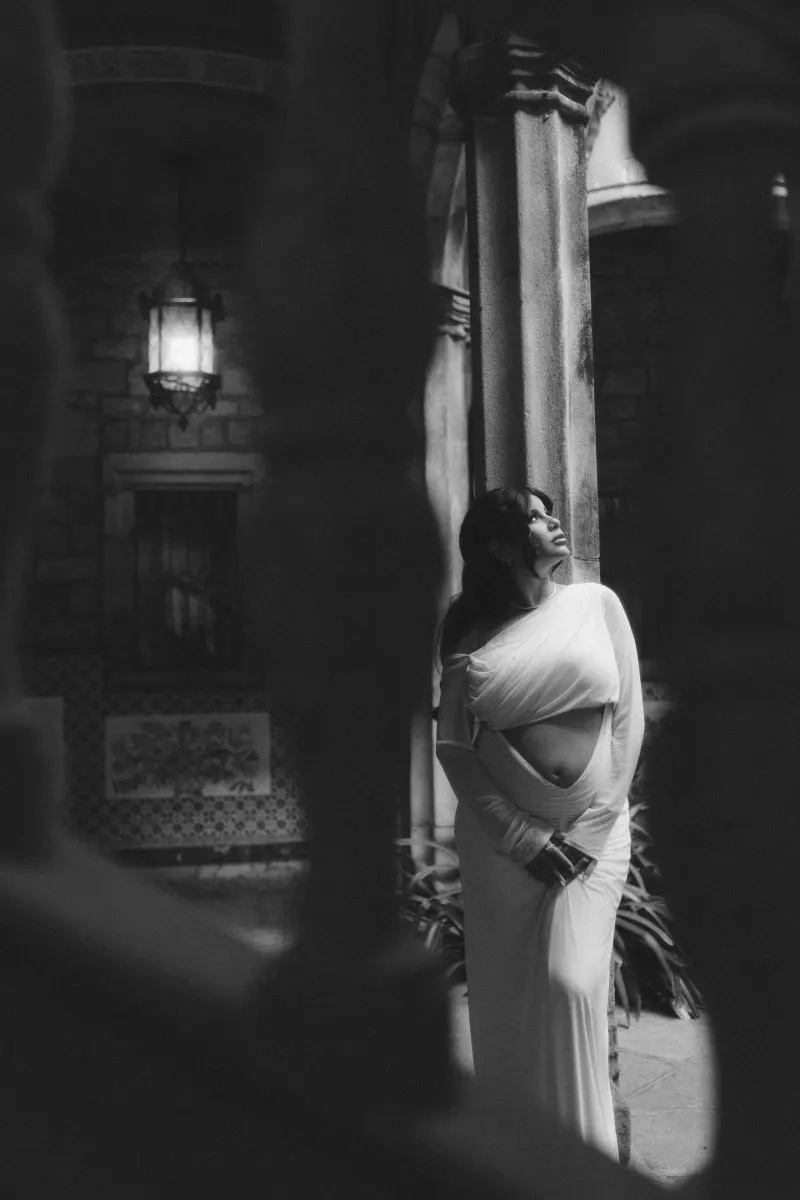 Pregnant woman in a white gown smiles softly while standing by a stone pillar during a Vacation Portraits session.