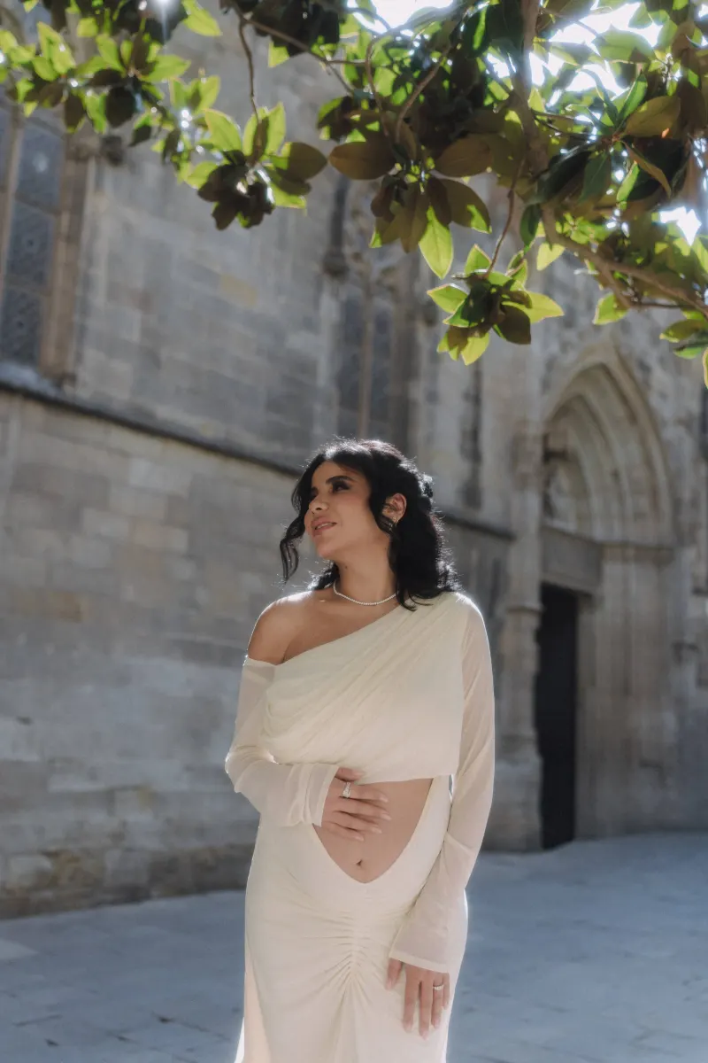 Pregnant woman in an elegant white dress smiles softly beneath leafy branches during a Vacation Portraits session.