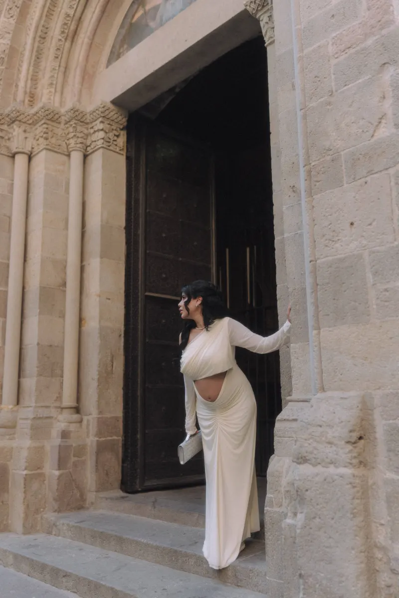 Radiant pregnant woman in a white gown pauses at a grand stone doorway during a Vacation Portraits photoshoot.