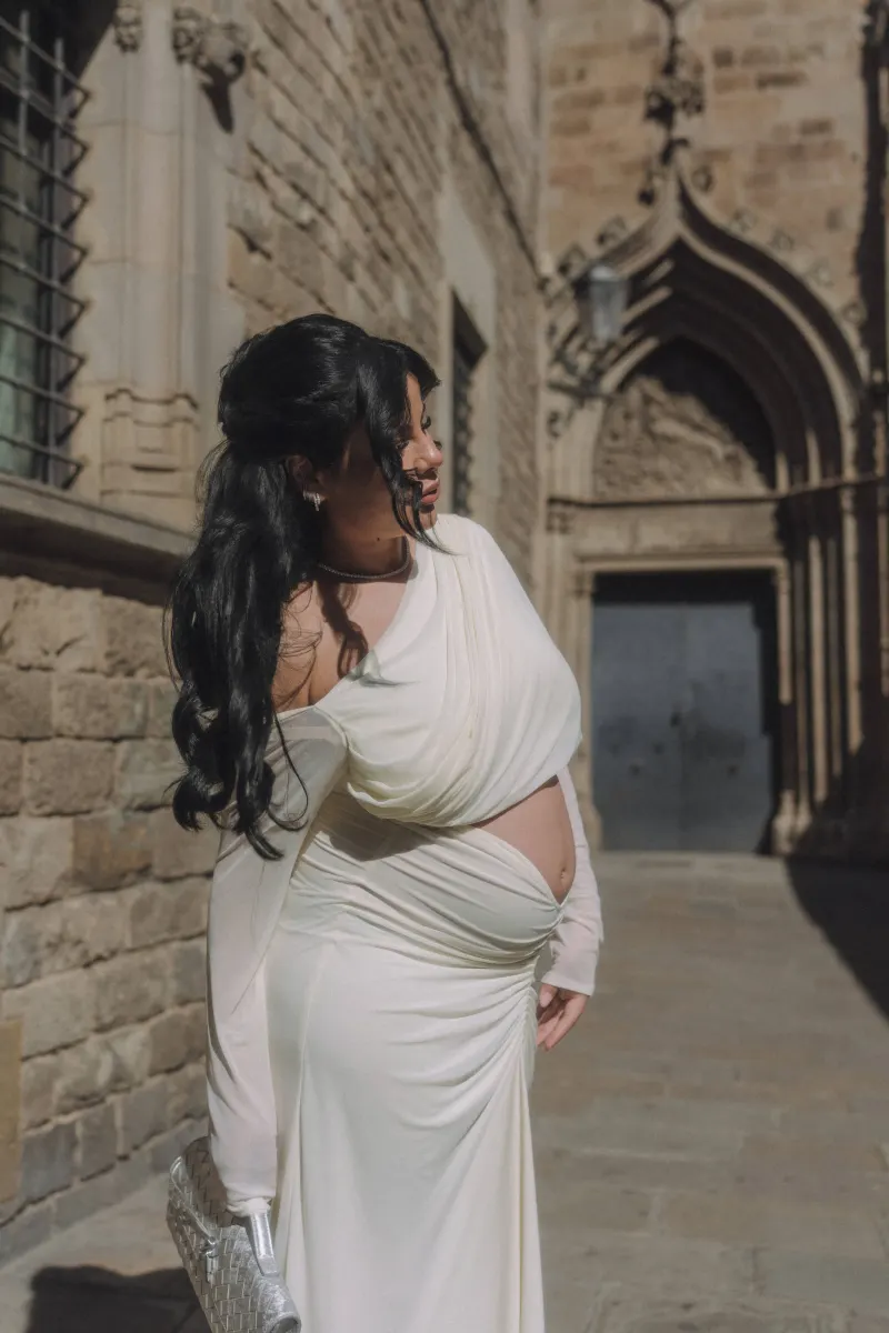 Radiant pregnant woman in a white gown smiles softly in a sunlit old city street during a Vacation Portraits session.