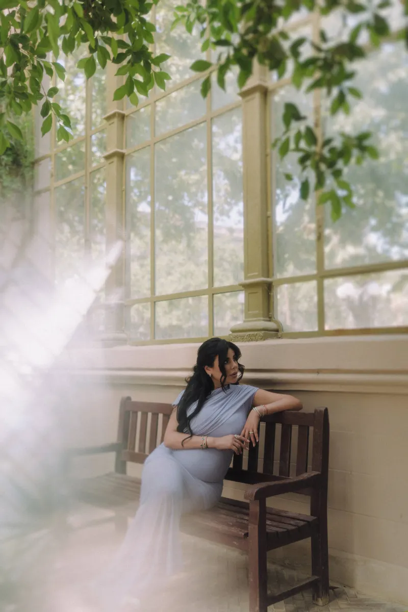 Relaxed expectant mother in a soft blue gown sits on a wooden bench, Vacation Portraits session in a sunlit conservatory.