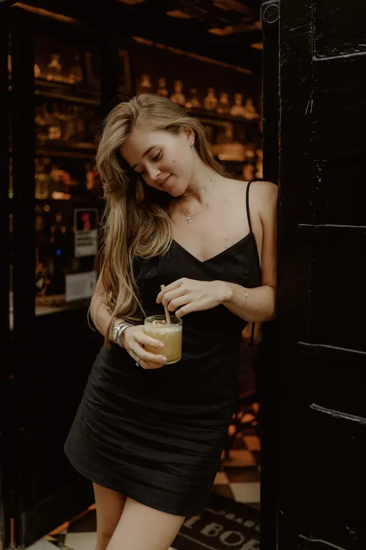 Relaxed woman in a black dress enjoys a cocktail at a cozy bar doorway during a Vacation Portraits photoshoot abroad.