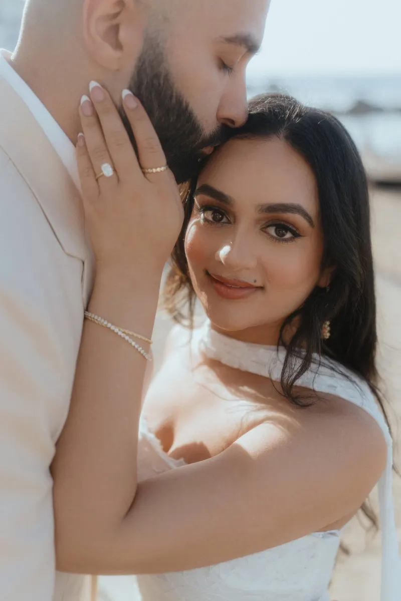Romantic beachside surprise proposal photoshoot, groom kisses bride’s forehead as she smiles, showing her engagement ring.