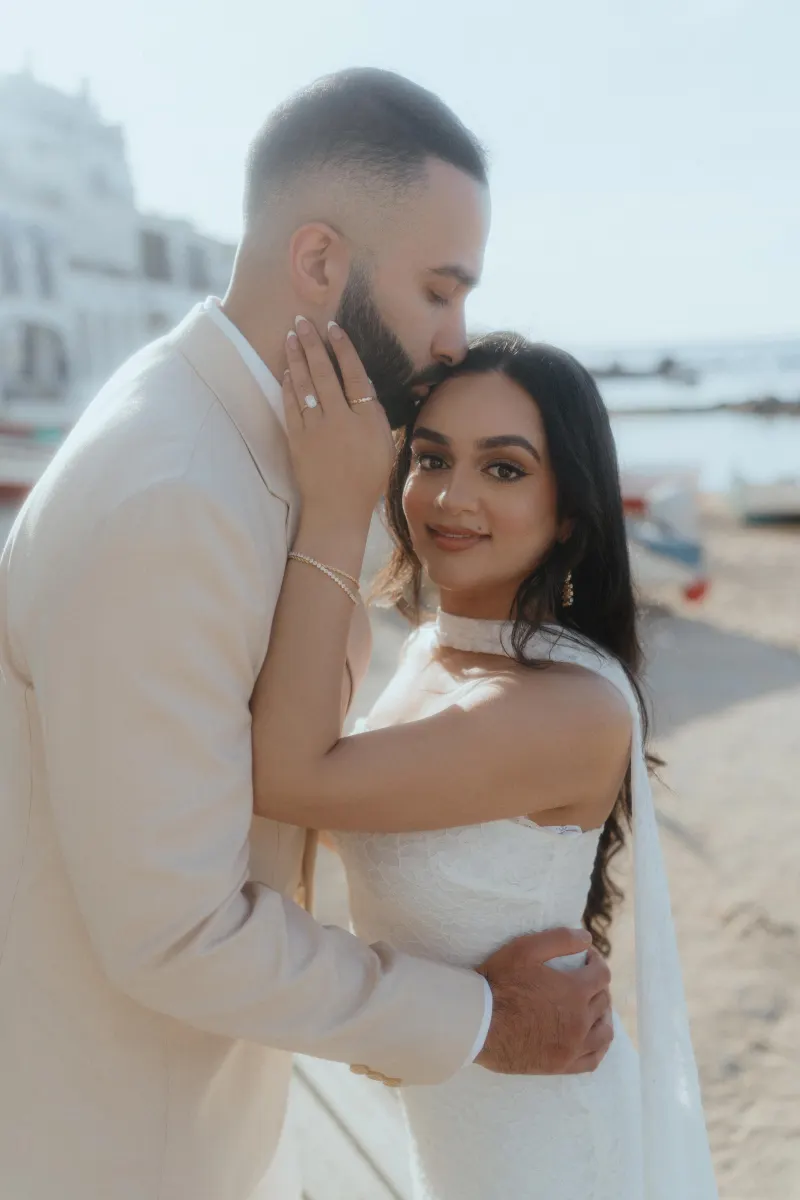 Romantic surprise proposal photoshoot by the beach, groom-to-be kisses fiancée’s forehead as she shows her ring.