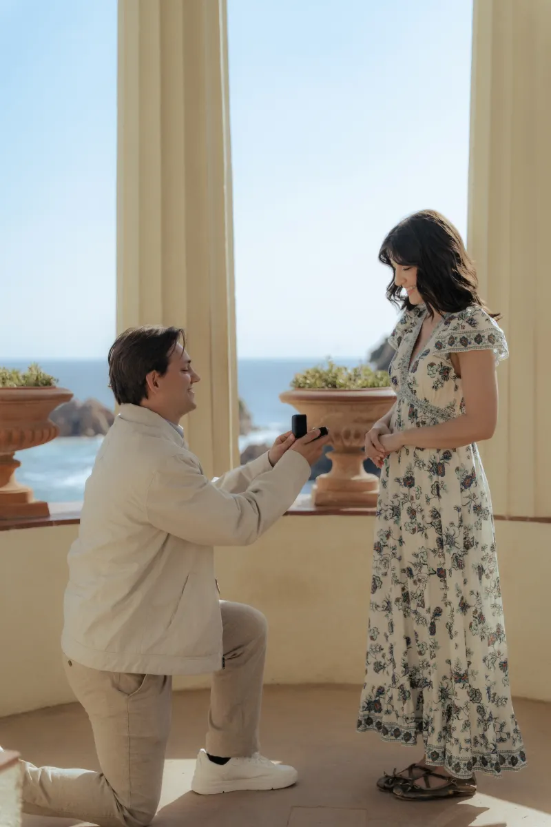 Romantic surprise proposal photoshoot by the ocean, capturing joy as he kneels with a ring and she smiles warmly.