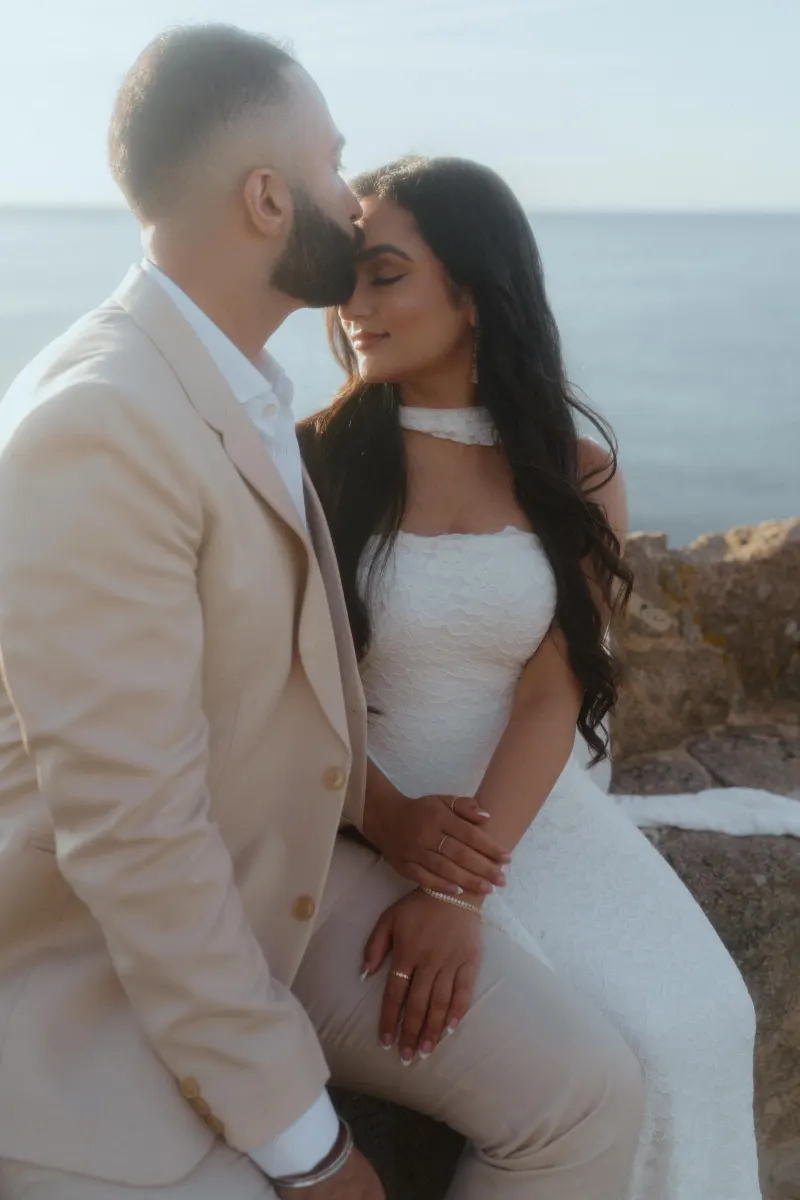 Romantic surprise proposal photoshoot by the ocean, groom kisses bride’s forehead as she smiles with eyes closed.