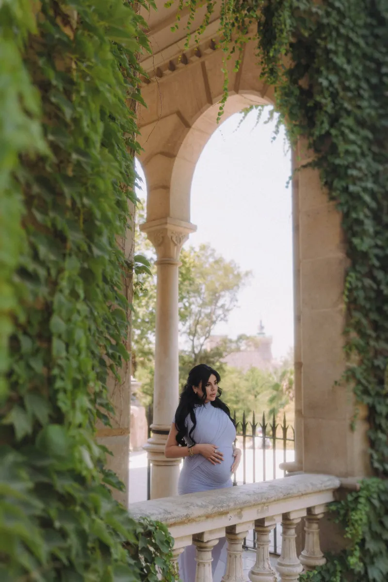 Serene expectant mother in a blue gown stands on a leafy balcony, Vacation Portraits session in Barcelona.