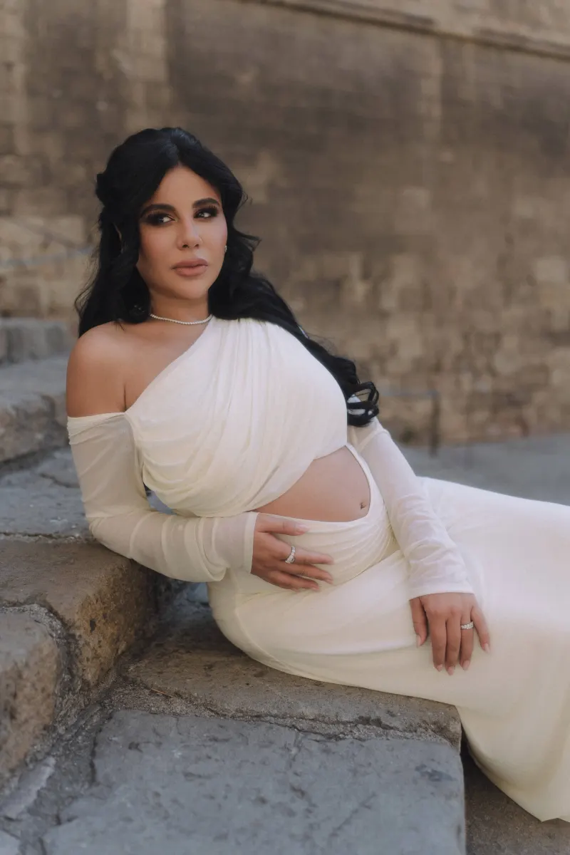 Serene expectant mother in a flowing white dress sits on old stone steps, captured during a Vacation Portraits session.
