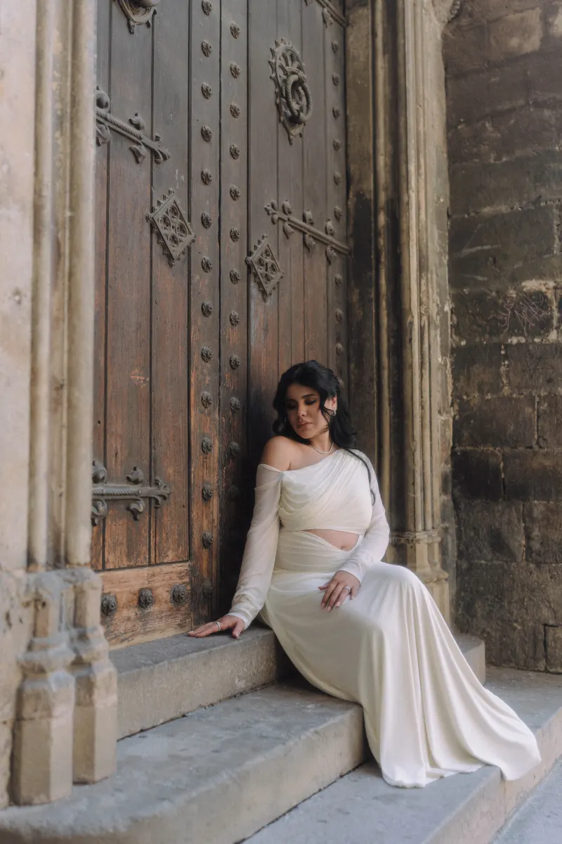Serene expectant mother in a white dress sits gracefully by an ornate wooden door during a Vacation Portraits session.