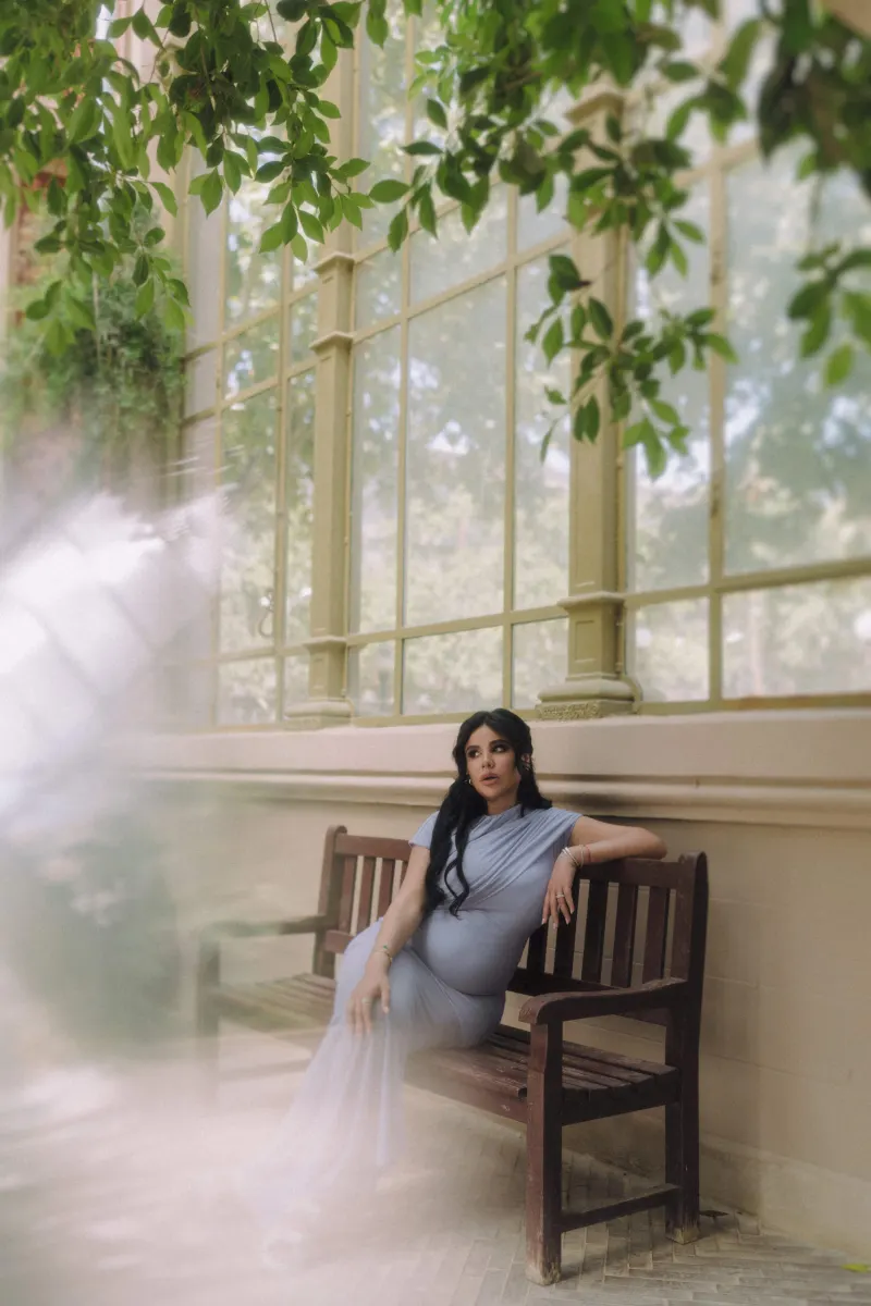 Serene mother-to-be in a flowing blue dress relaxes on a bench, Vacation Portraits photoshoot in a leafy conservatory.