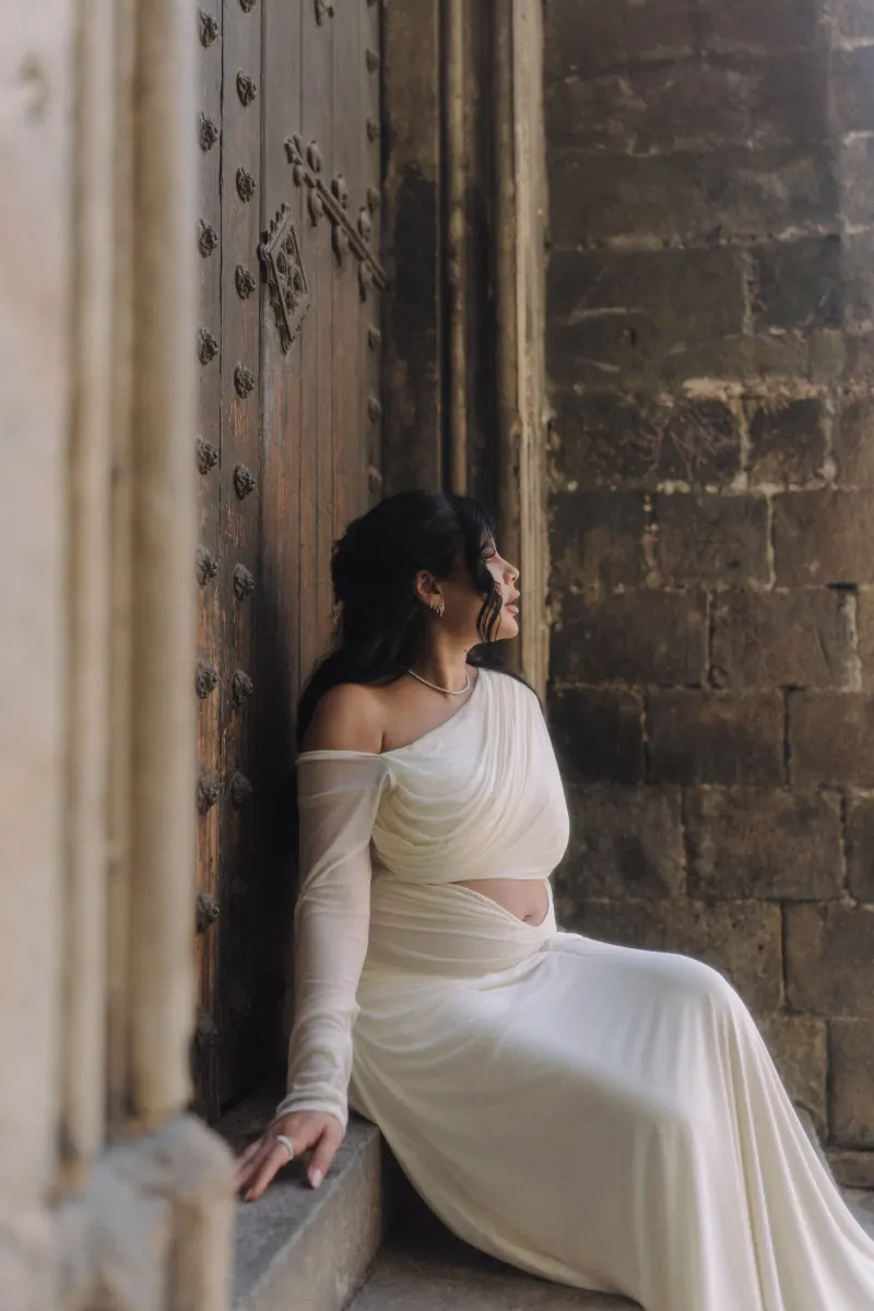Serene pregnant woman in a flowing white dress rests by an ancient doorway during a Vacation Portraits session.