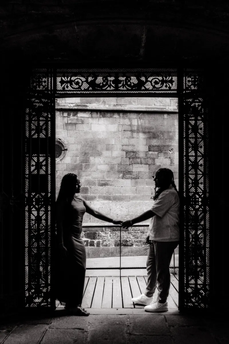 Silhouetted couple holds hands through ornate gates, sharing a quiet moment during a surprise proposal in Barcelona.