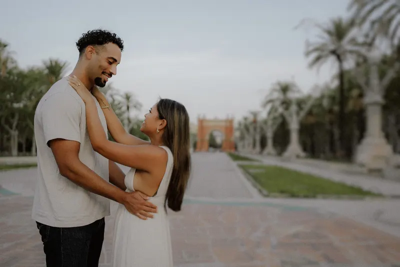 Smiling couple embracing during an engagement session, surrounded by palm trees and historic architecture.