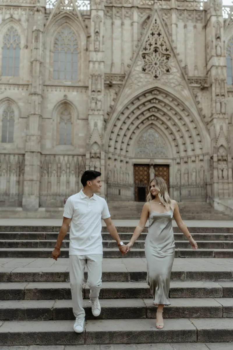 Smiling couple holding hands on cathedral steps in Barcelona during a Vacation Portraits photoshoot.