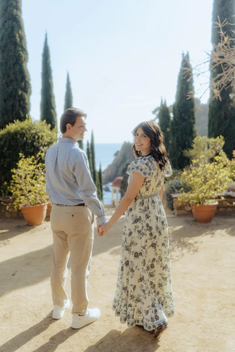 Smiling couple holds hands on a sunlit garden path, capturing a candid moment from their surprise proposal photoshoot.