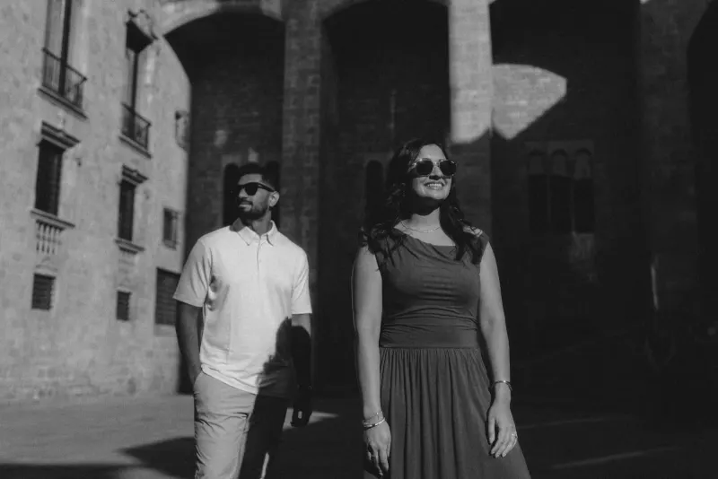 Smiling couple in sunglasses enjoys a sunlit stroll during their couples & engagements photoshoot in a historic courtyard.