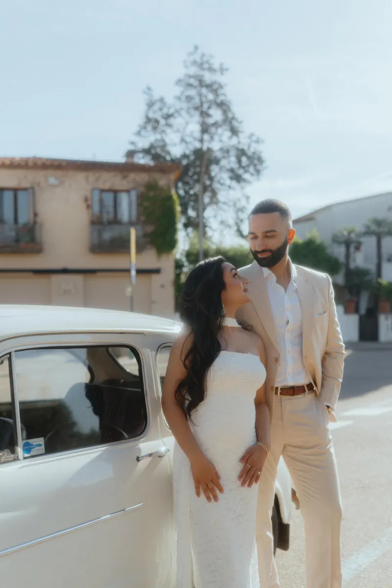 Smiling couple shares a romantic moment by a vintage car after their surprise proposal photoshoot in a charming plaza.