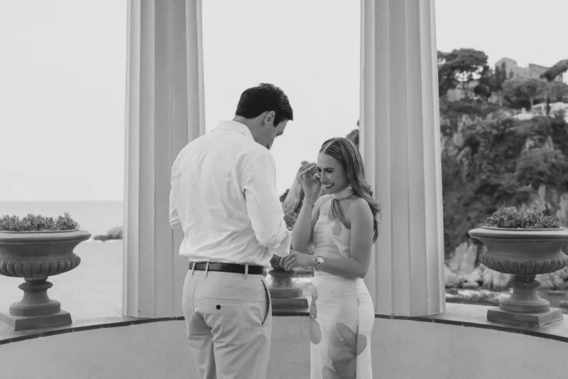 Smiling couple shares an emotional moment during a seaside surprise proposal photoshoot at an elegant pavilion.
