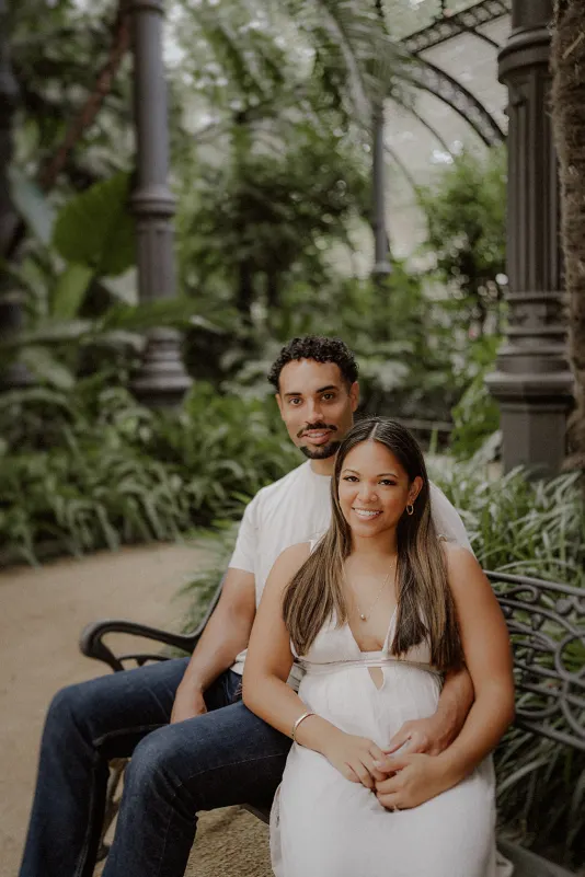 Smiling couple sits close together on a garden bench, sharing a relaxed moment during their engagement photoshoot.