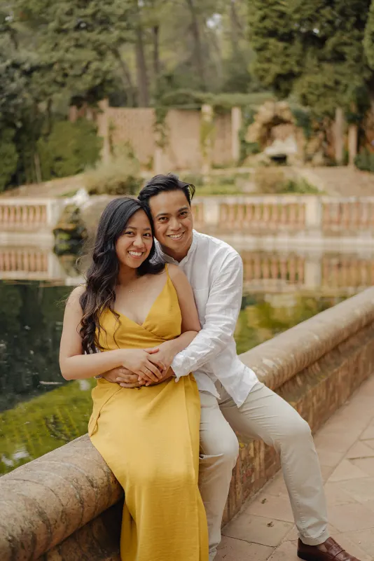 Smiling couple sits together by a peaceful pond, sharing a warm embrace during their couples & engagements photoshoot.