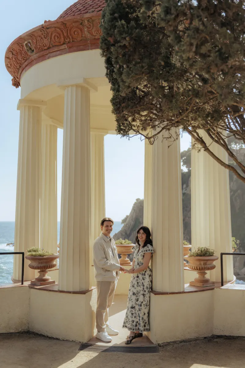 Smiling couple stands hand in hand beneath seaside columns, captured during a surprise proposal photography session.
