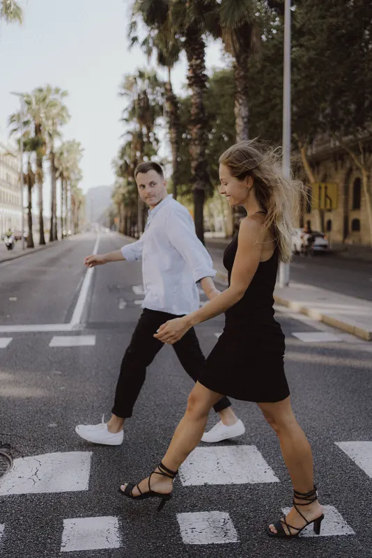 Smiling couple walks hand in hand across a palm-lined street during a couples & engagements photoshoot.