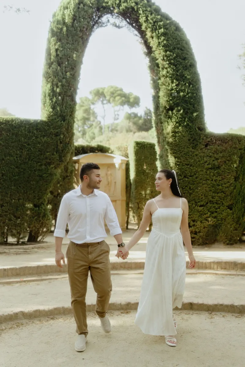 Smiling couple walks hand in hand through a sun-dappled garden arch during a couples & engagements photography session.