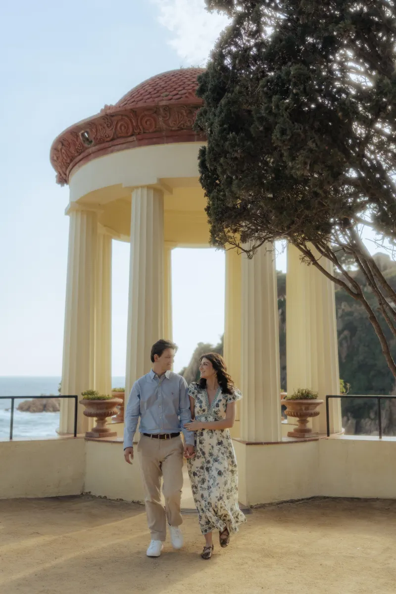 Smiling couple walks together near a seaside pavilion, capturing their engagement during a surprise proposal photoshoot.